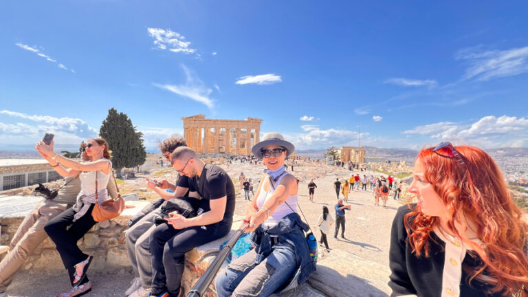 Moon Suk mit Selfie-Stick vor der Akropolis, umgeben von Menschen; Athen-Reiseführer-Szene.