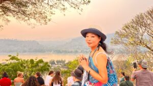 MOON SUK schaut bei Sonnenuntergang in Luang Prabang, Kambodscha, in die Kamera, umgeben von Menschen, die den Ausblick genießen. Ein symbolisches Bild ihrer Reisen als Künstlerin.