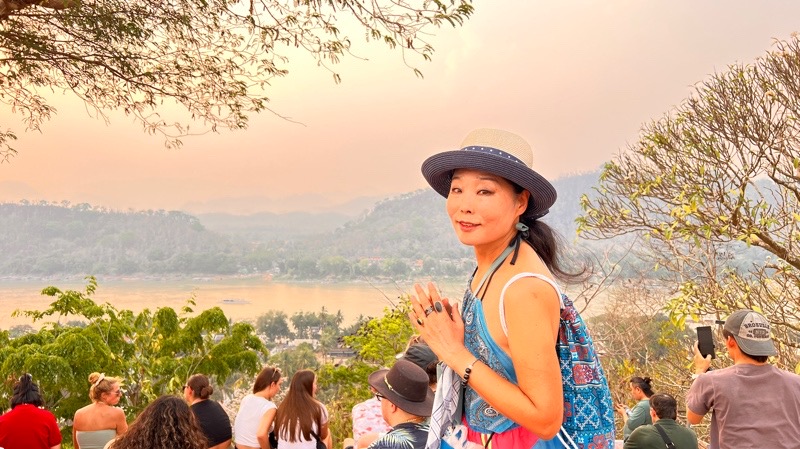 MOON SUK schaut bei Sonnenuntergang in Luang Prabang, Kambodscha, in die Kamera, umgeben von Menschen, die den Ausblick genießen. Ein symbolisches Bild ihrer Reisen als Künstlerin.