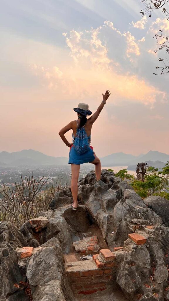 MOON SUK allein auf einem Felsen vor Sonnenuntergang in Luang Prabang, Laos, mit dem Rücken zur Sonne, symbolisiert Freiheit und künstlerische Reflexion.