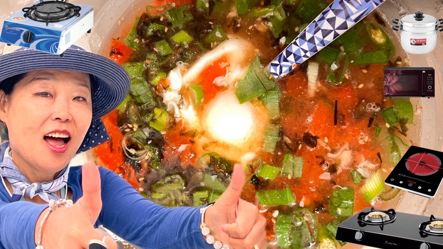 Moon Suk mit zwei Daumen hoch präsentiert perfekt pochiertes Ei und Nudeln aus dem Wasserkocher, umgeben von Küchenutensilien und einem Herd im Hintergrund.