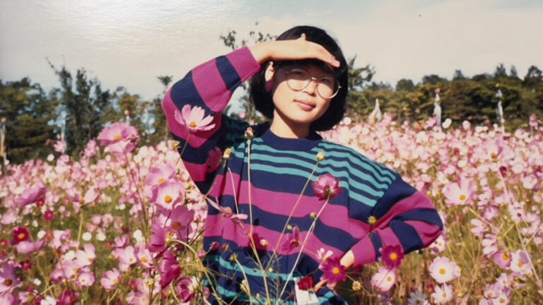 Moon Suk mit Brille in einem pink-grünen Pullover, posiert auf einem Kosmos-Blumenfeld in Korea, eine Hand zum Schutz vor der Sonne an der Stirn.